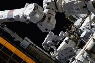 two spacewalking nasa astronauts work near the international space station's solar arrays, with the orbiting lab's canadarm2 robotic arm visible in the foreground.