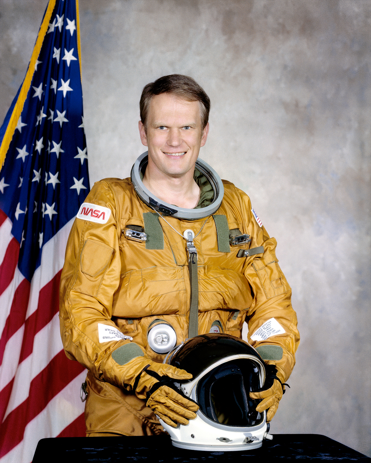 an astronaut in an orange flight suit poses in front of the American flag.