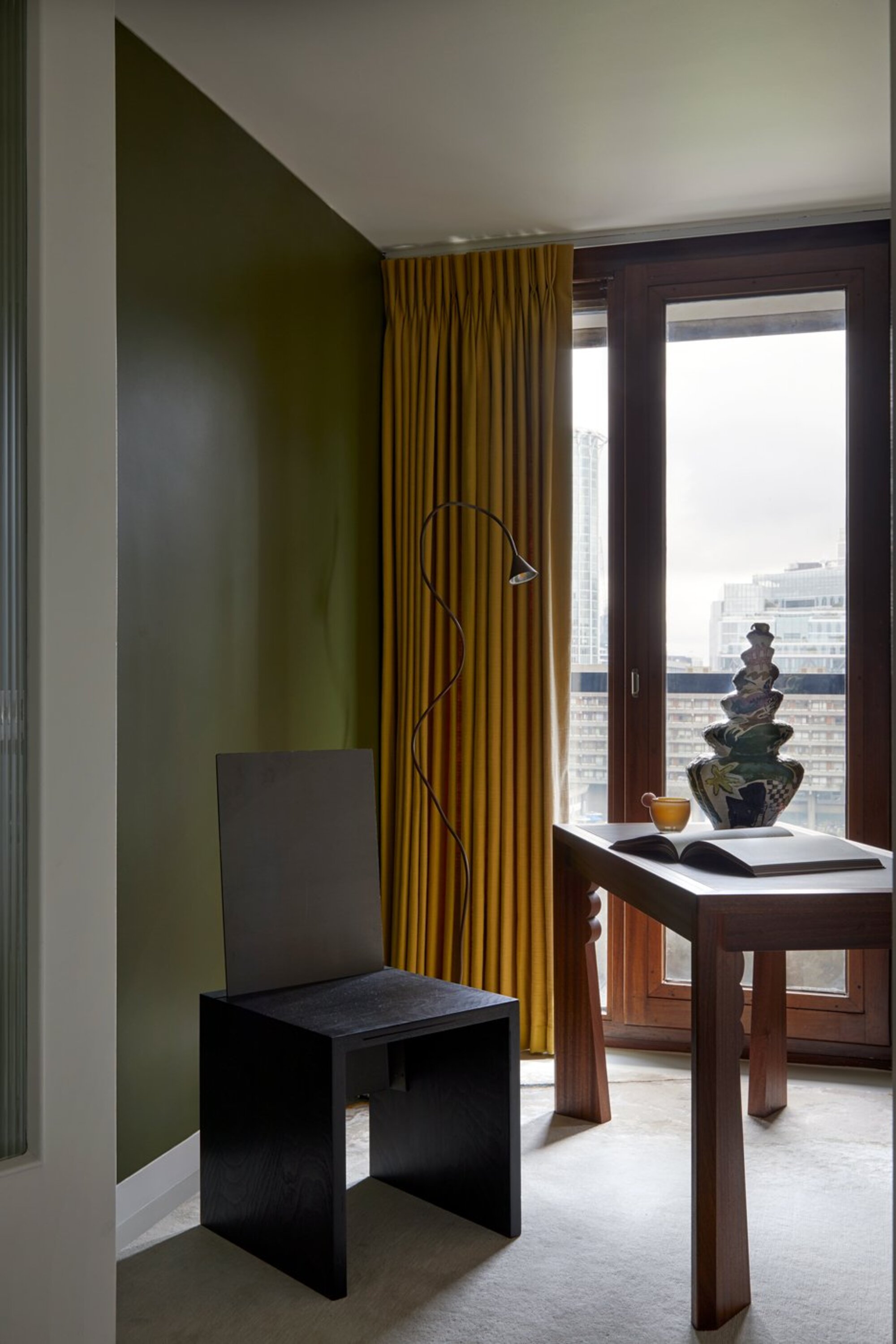 home office with green wall, mustard yellow curtains, a black sculptural chair next to a timber desk with statue and book