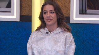 Dee sitting at the table in a white hoodie with grey tinges at the Big Brother dining room.
