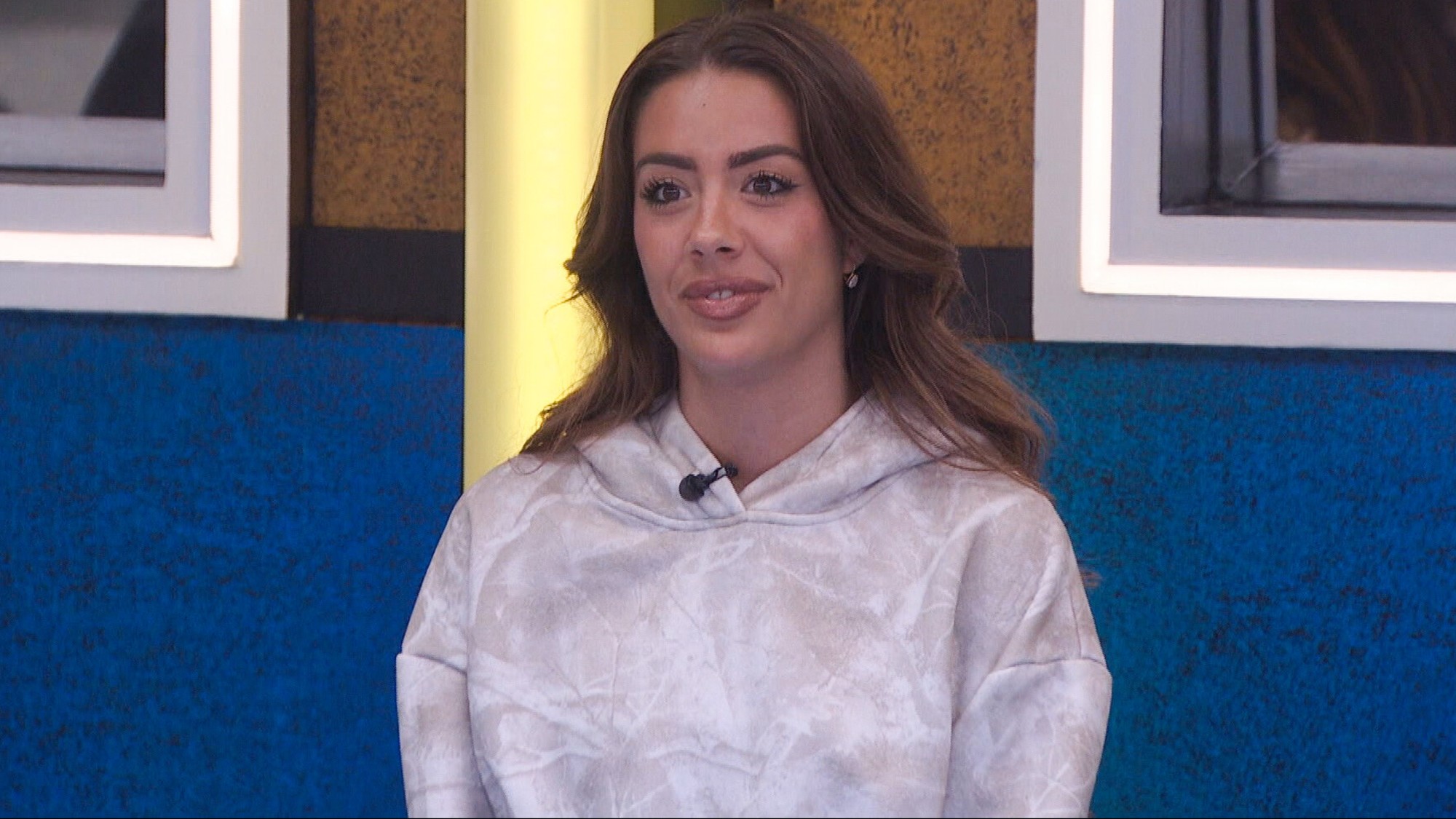 Dee sitting at the table in a white hoodie with grey tinges at the Big Brother dining room. 