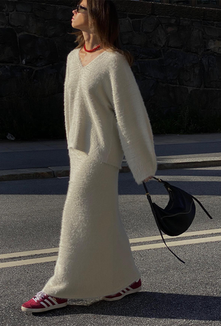 what to wear in 30-degree temperature is shown in a photo of a woman walking in the street wearing black sunglasses, a red necklace, an oversize white cashmere v-neck sweater with a matching knit maxi skirt, red sneakers, and a black crescent bag