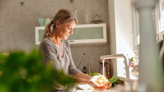 Woman smiling and washing oranges in front of kitchen window with sun coming through