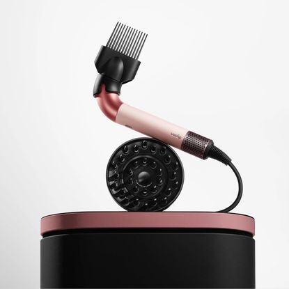 Dyson supersonic r blow dryer with wide-tooth and diffuser attachment sitting on pink and black carrying case against grey gradient background 