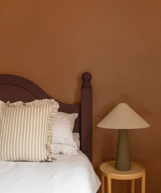 bedroom with dark orange wall, dark red bed frame and white bedding with neutral striped cushion and small table with modern lamp