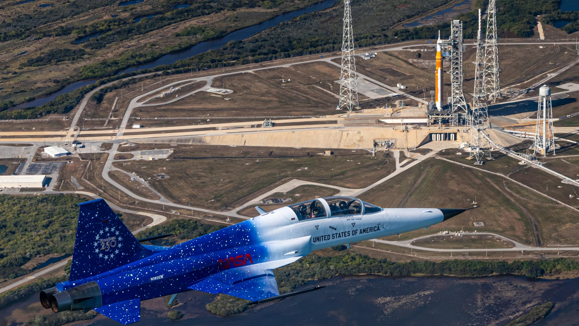 A blue and white jet flies over a launch complex where an orange and white rocket sits on a launch pad