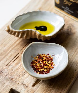 Two shell shaped bowls, one with oil and balsamic in it and one with chilli flakes on it, on a light wooden board