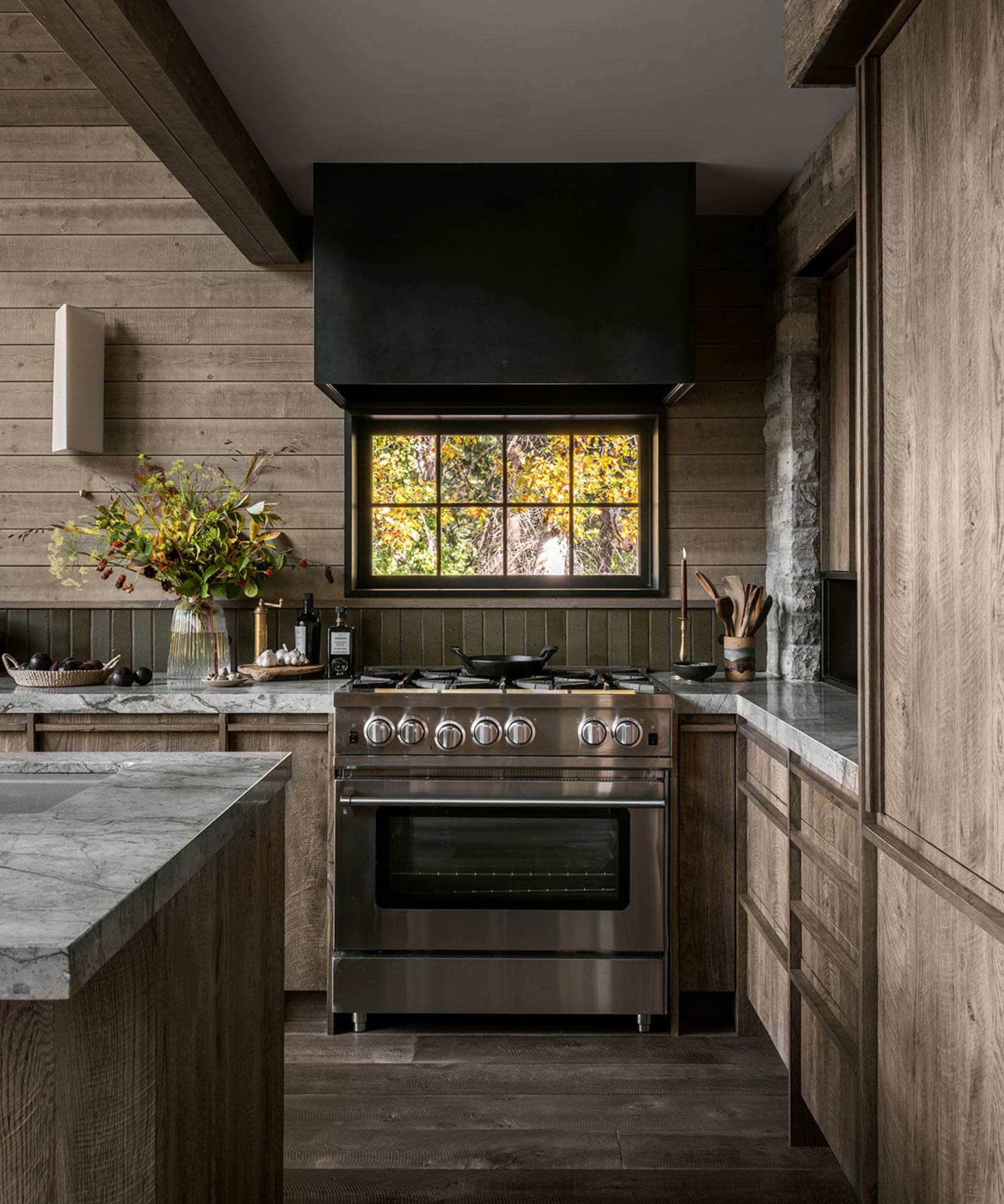 A cabin kitchen with wooden cabinets, marble countertops, and a mixed material backsplash featuring green tiles and wood panels