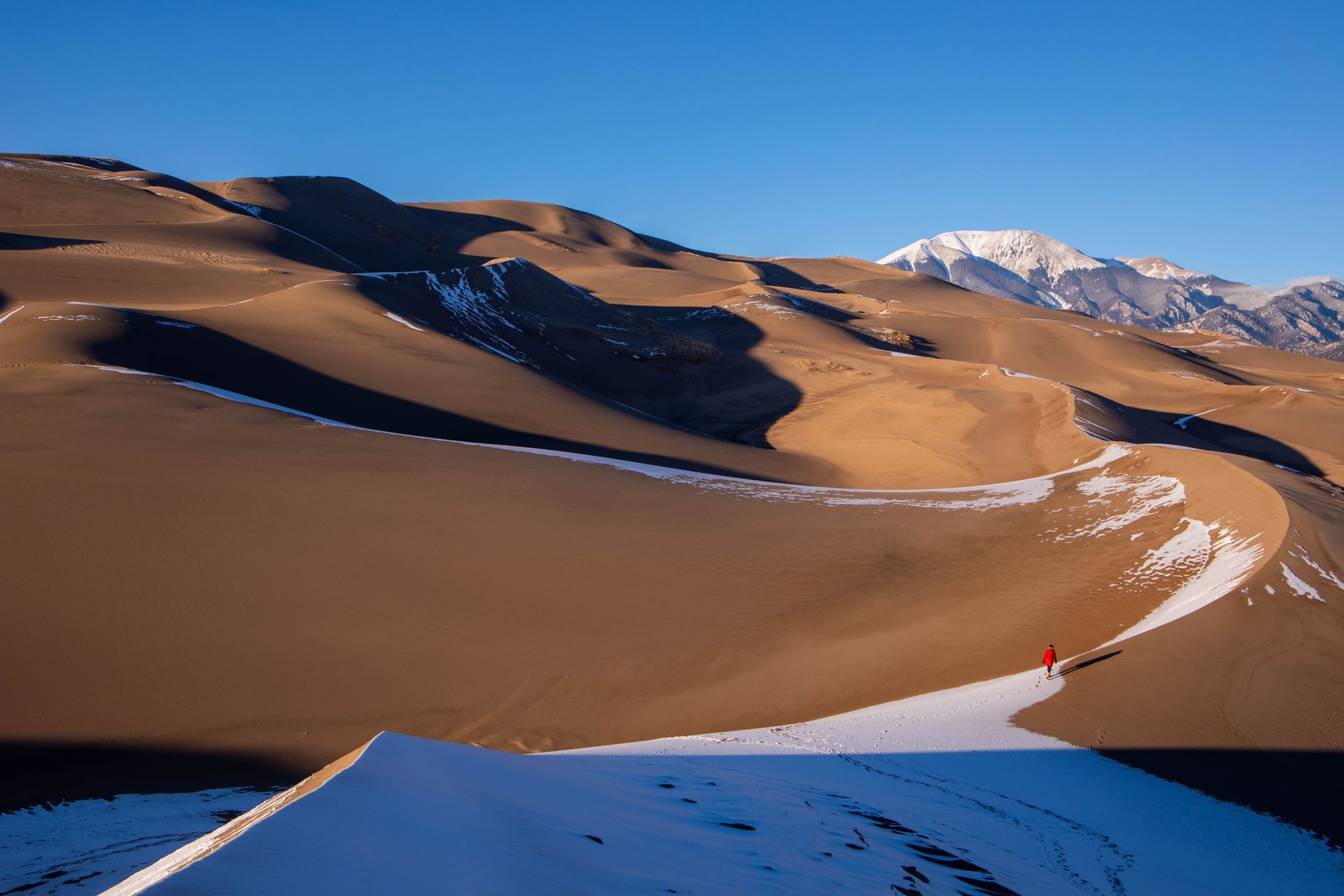 The sun hitting Great Sand Dunes National Park with a person walking and mountains in the background