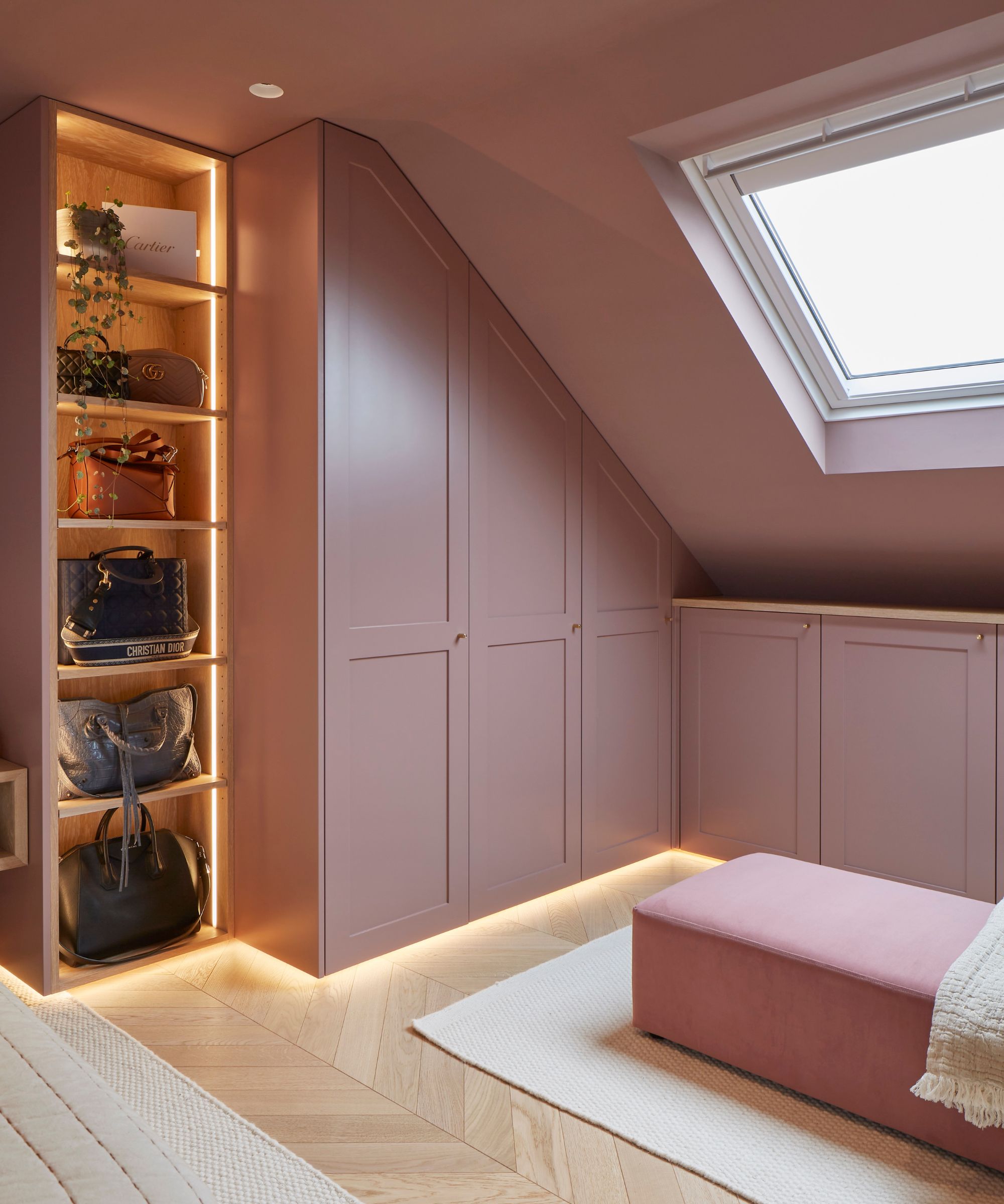 A softly lit loft dressing room with sloped ceilings and a large rectangular skylight bringing in natural light. The cabinetry, walls and built-in wardrobes are painted in a muted dusty pink tone, creating a cohesive, cocooning feel. Along one wall, low fitted cupboards run beneath the slanted ceiling, with subtle under-cabinet lighting casting a warm glow along the floor.