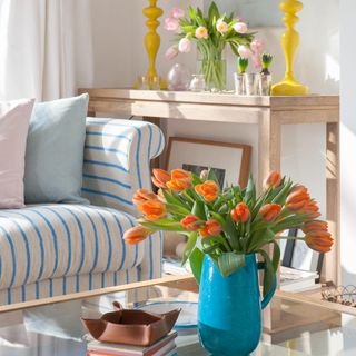 A living room with a blue striped sofa and a glass coffee table with a vase of orange tulips