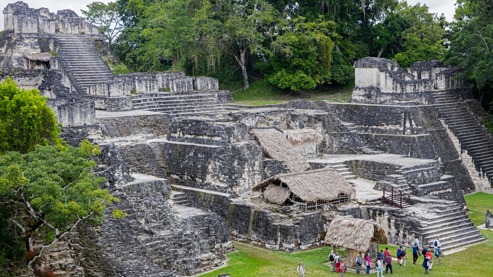 North Acropolis of Tikal. Three smaller step pyramids side by side, and then at the top of these there is a larger step pyramid in the center. Each has a wide staircase leading to the top. There is a small grass hut at the very bottom, and two more grass buildings on the middle step pyramid. There is a large group of people (possibly tourists) at the bottom.