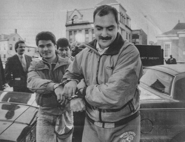Two men in handcuffs, one with a mustache, walk away from a police car and into a building.