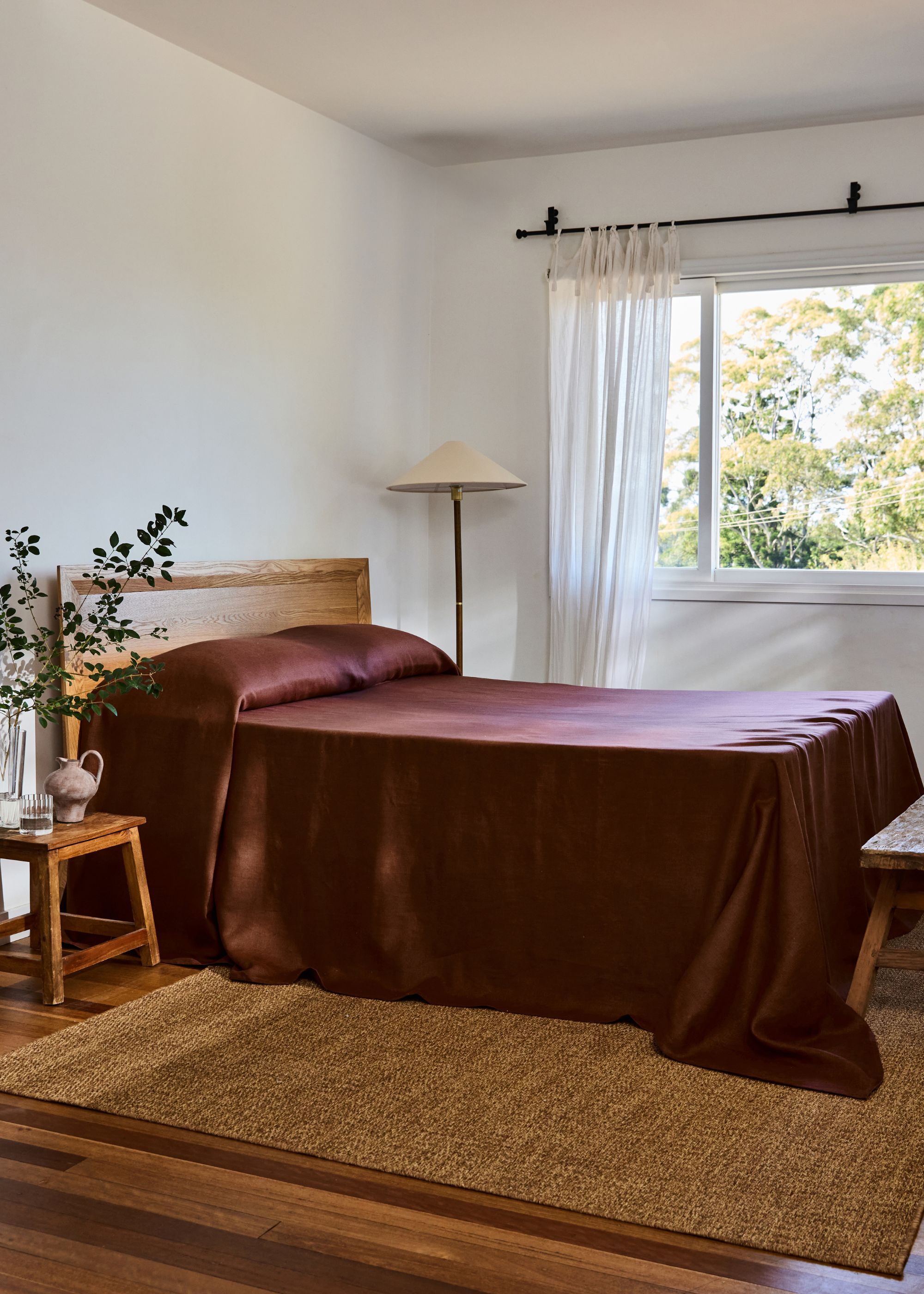 A bedroom with a large window, white curtains, a maroon satin bed cover, and a floor lamp