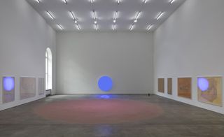 Six paintings hang in a gallery with a blue illuminated circle at one end of the room