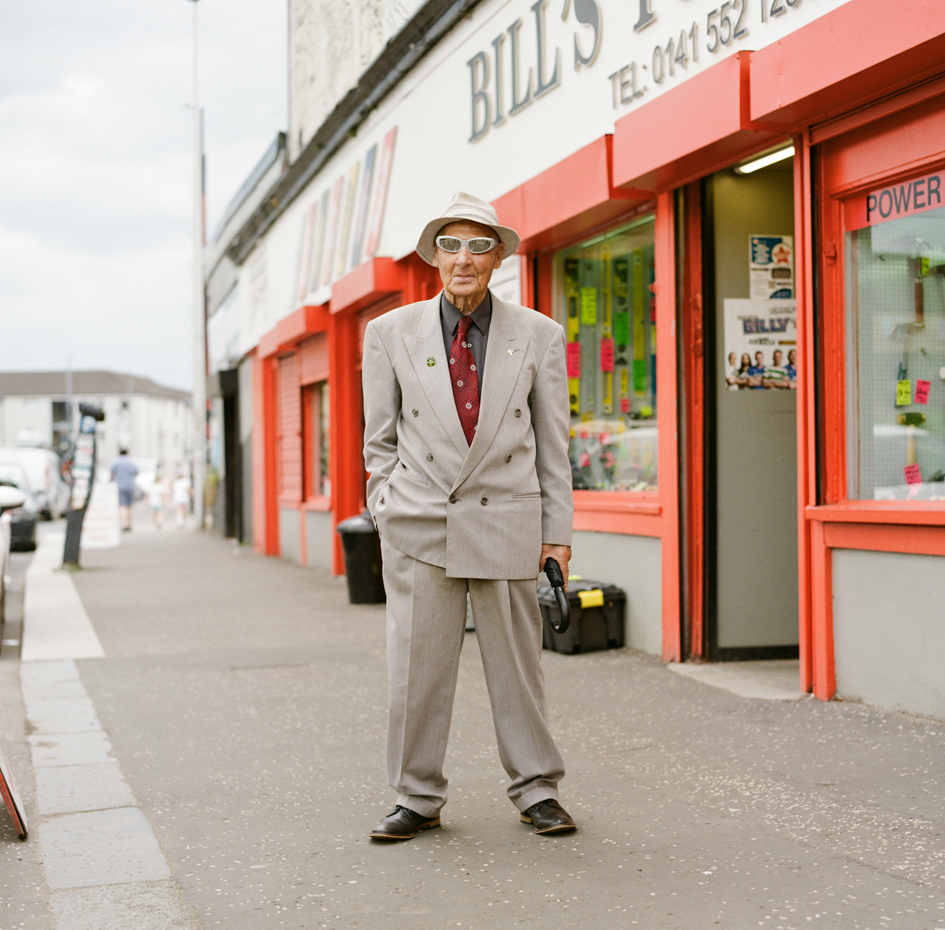 Elderly man in a gray suit and hat, with sunglasses, stands confidently in front of a colorful shop with red accents on a cloudy day