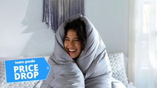 A woman laughs while sitting on a bed wrapped up in a grey Layla Down Alternative Comforter; a blue price drop sales badge is overlaid on the bottom left hand corner