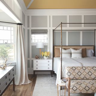 how to panel a wall, grey and yellow bedroom with paneling, four poster metal bed, bay window, yellow ceiling, small sofa at foot of bed, natural bedding