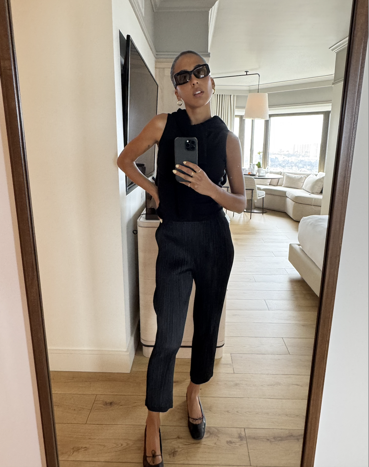Madison Utendahl posing in mirror wearing black pleats please issey miyake outfit, sunglasses, and ballet flats