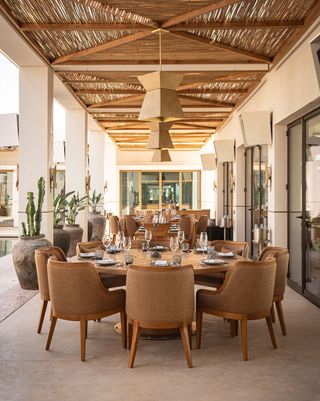 In an outdoor patio, multiple round dining tables and matching chairs in wood stand underneath a rattan ceiling decorated with pendant lanterns.