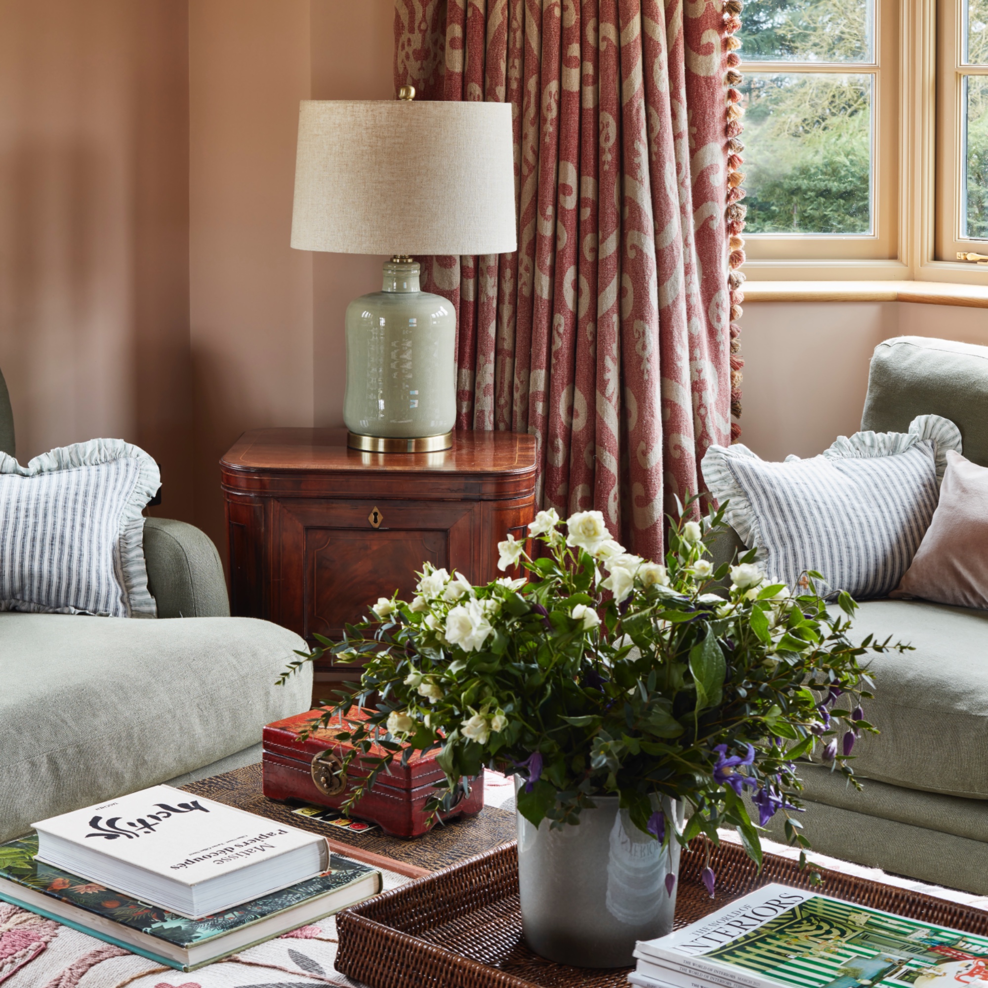 cosy living room with matching green sofas, footstool with tray and flowers, table lamp, patterned curtains