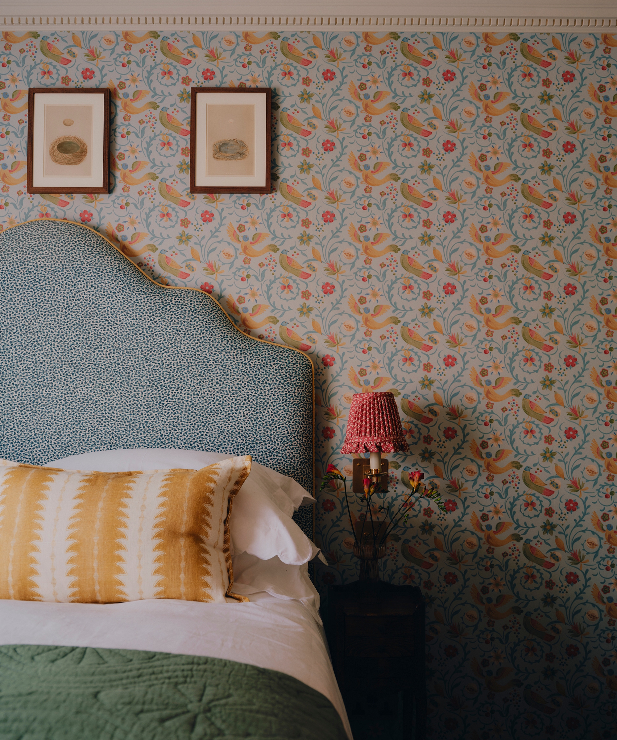 A bedroom with colorful botanical wallpaper, a blue patterned headboard and a red pleated floral wall lampshade.