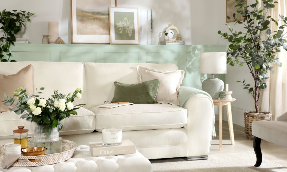 A cream upholstered sofa with green velvet and frilled cushions in a soft white living room with sage green panelling, eucalyptus branches and white rose arrangement