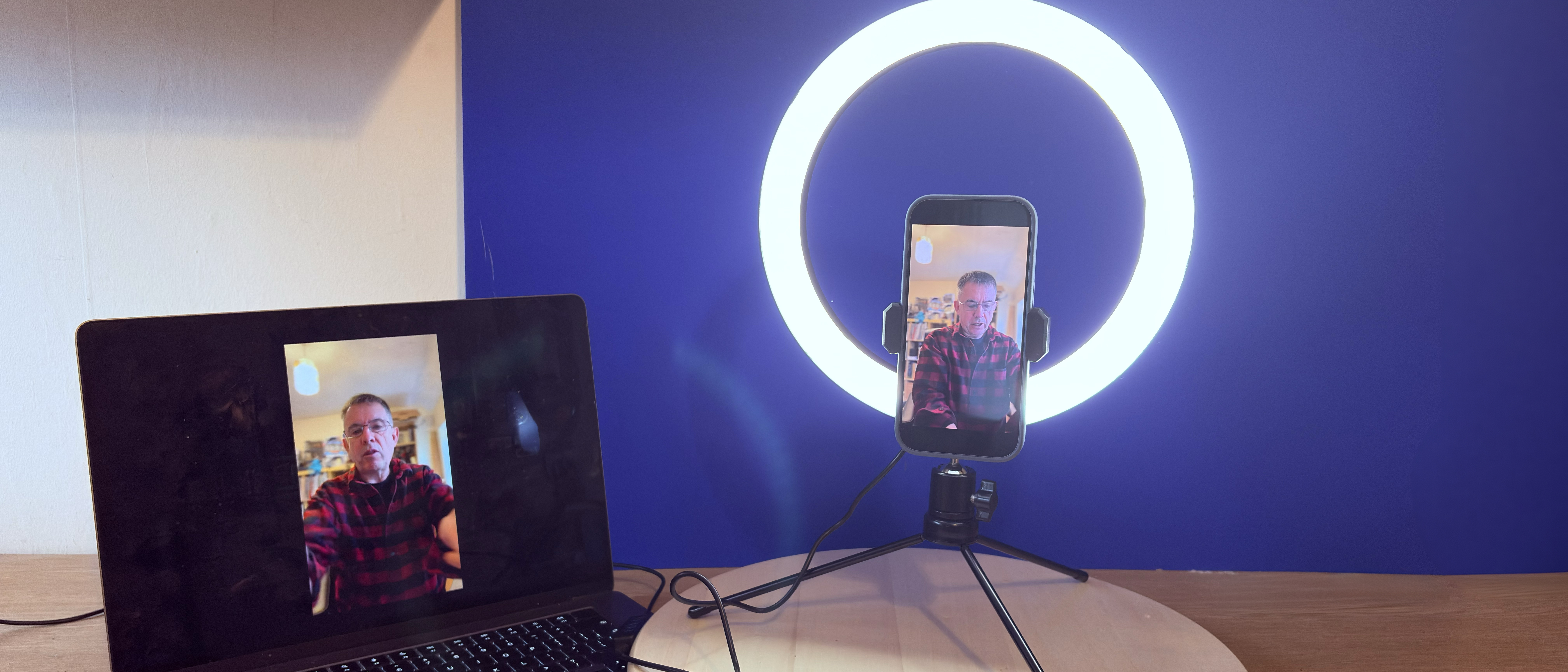 Shot of the AiXPi 10&rdquo; Ring Light holding an iPhone 17. The feed from the iPhone&rsquo;s camera is being displayed on a MacBook. A hand holds the LED&rsquo;s control unit.