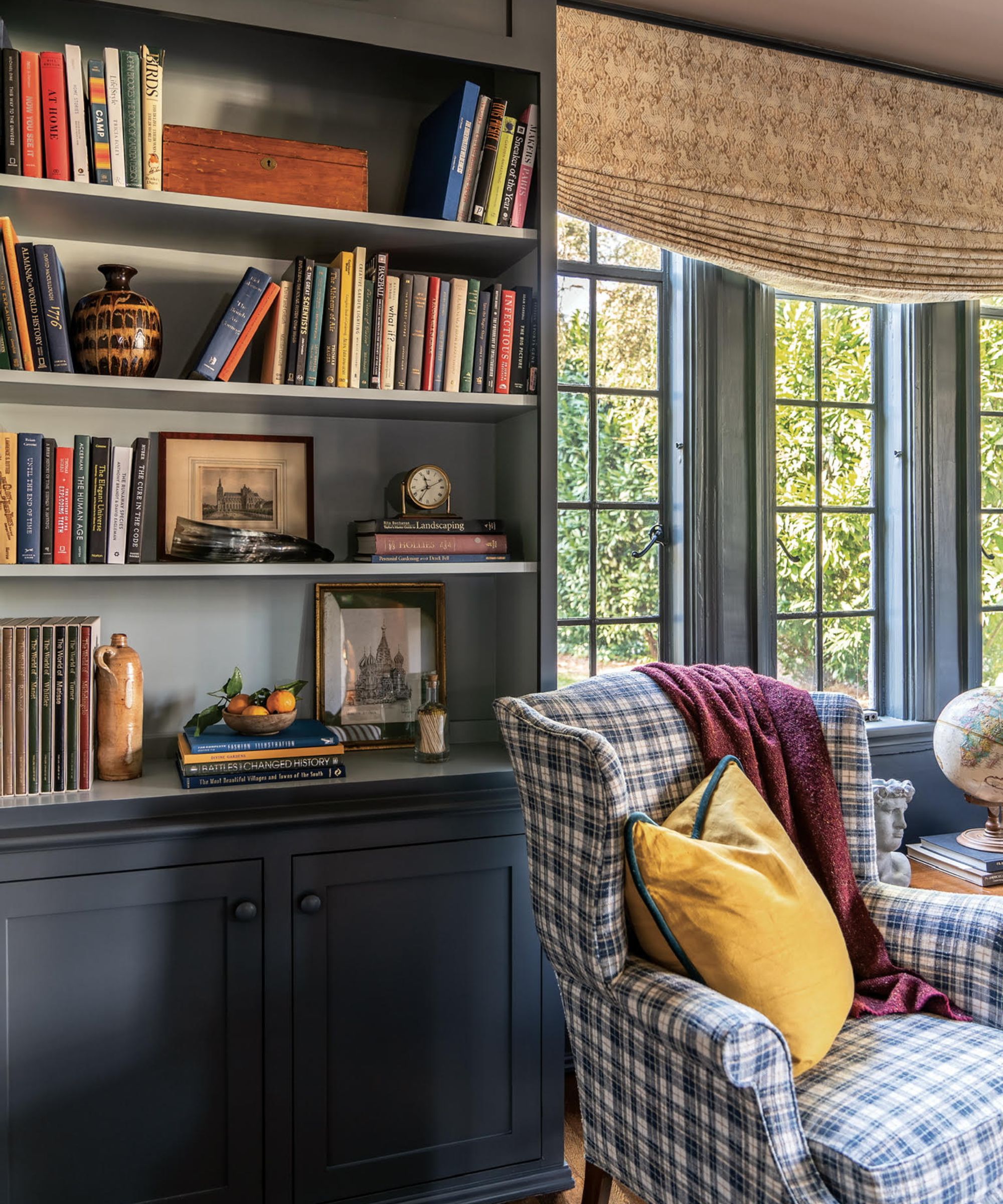 Living room painted bookshelf and windows in Flint by Benjamin Moore, a plaid blue armchair with a yellow cushion, and bookshelves filled with books and decor.
