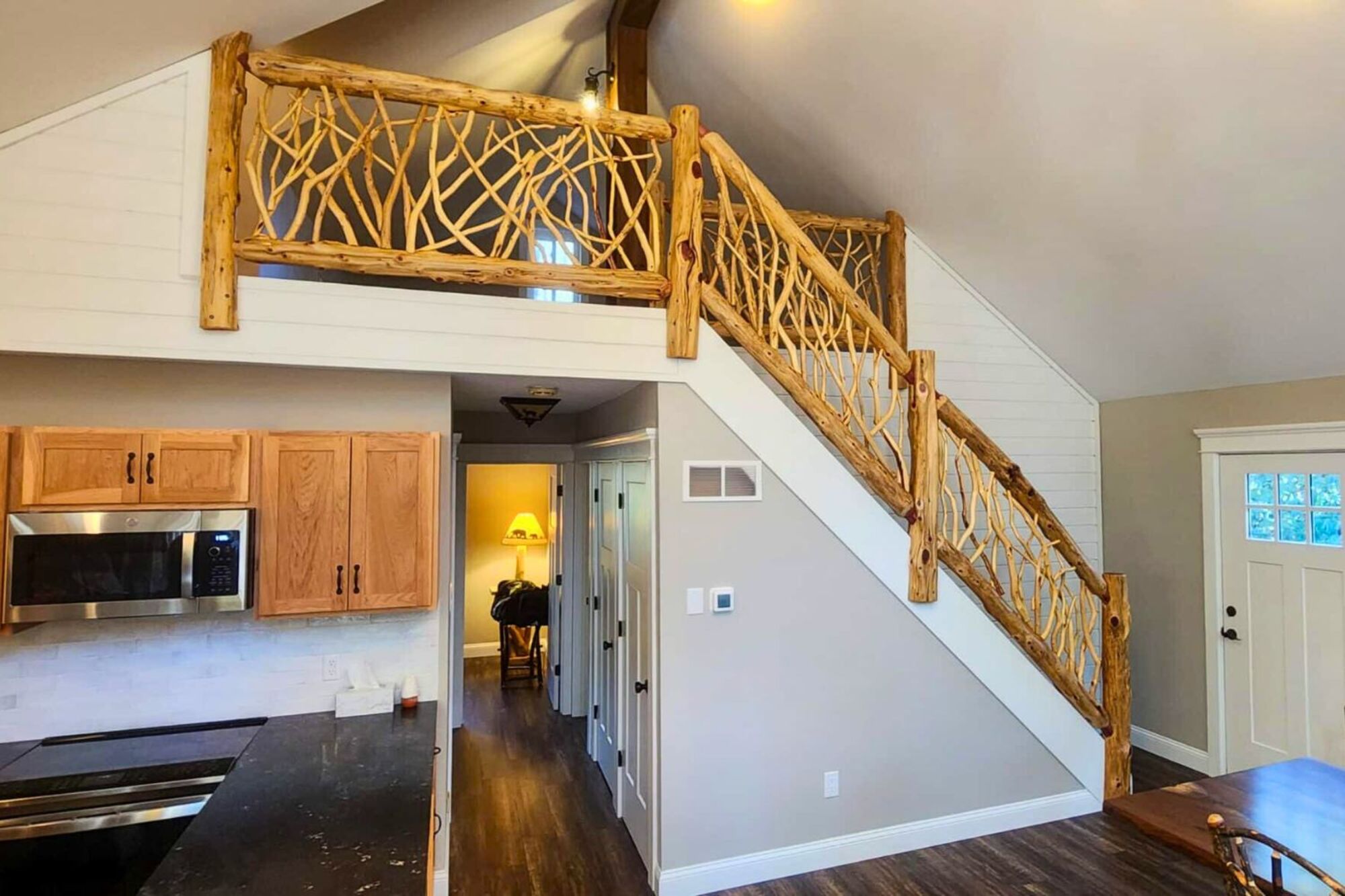 A studio with a small kitchen with wooden cabinetry and a set of stairs leading to a loft with an abstract, artisanal wooden ballustrade
