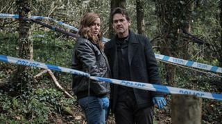 Eve Myles as Jeanette Kilburn and Dougray Scott as Lou Stanley in dark jackets stand near police tape in woodland in The Crow Girl.