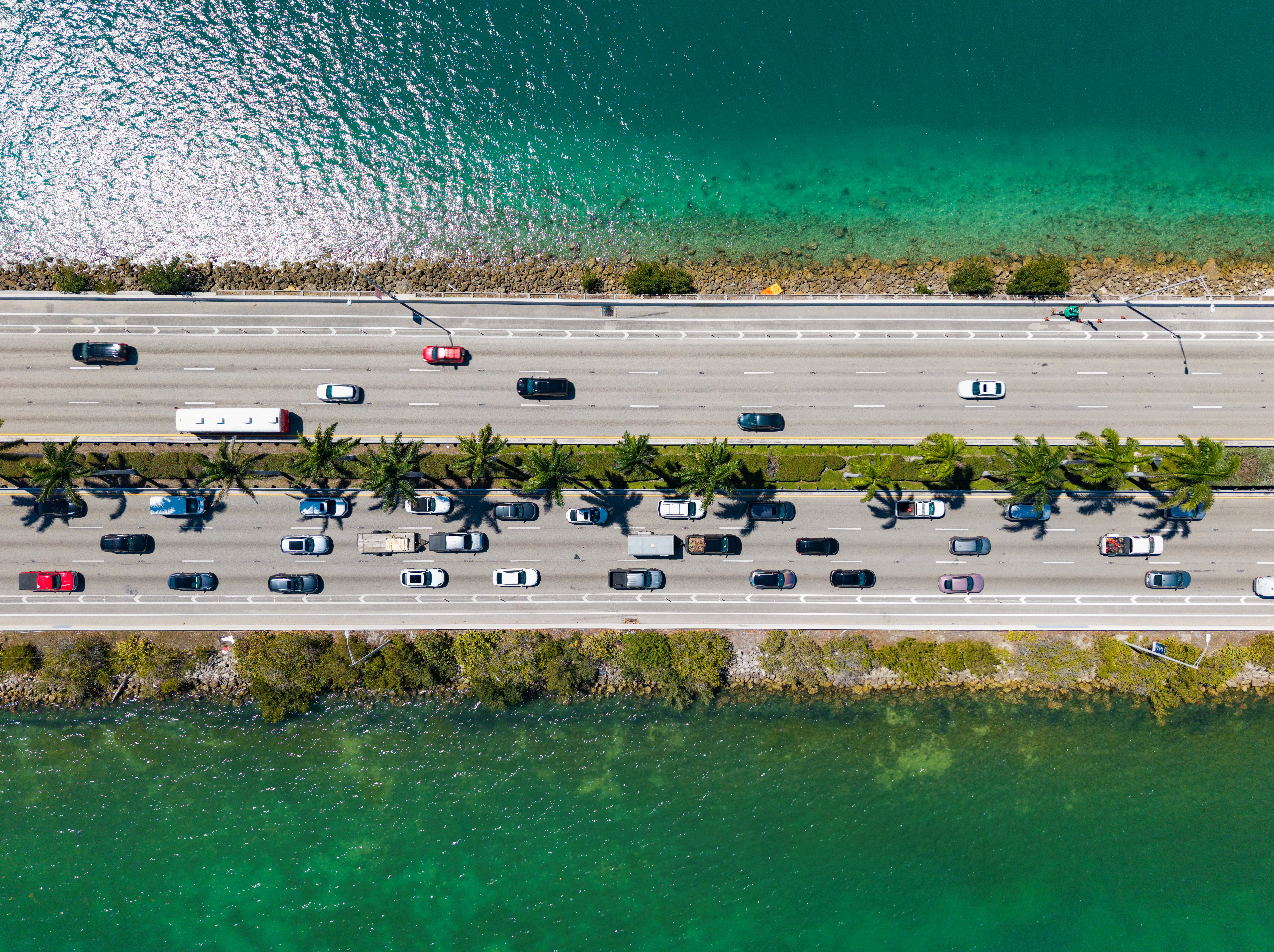 An aerial view of a highway surrounded, on either side, by green-to-blue Caribbean sea on a sunny day.