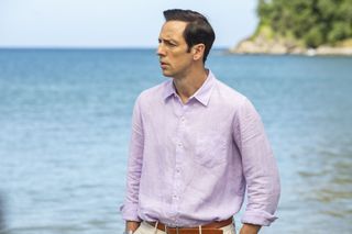 Death In Paradise season 13 episode 8: DI Neville Parker (Ralf Little) stands on the beach with the ocean behind him, looking pensive, with his hands in his pockets