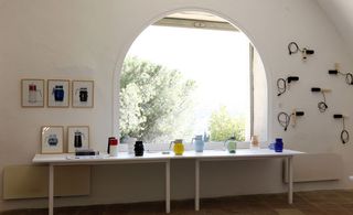 White table with variations of colourful electric kettles on display against a white wall with art displayed and an arch opening with a view of the outside