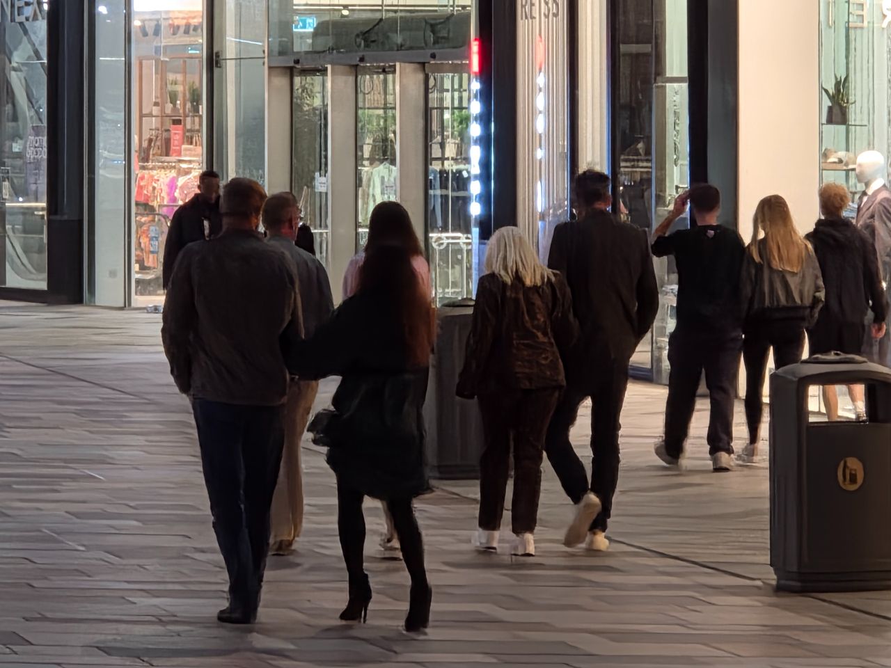 Close telephoto view of people walking past brightly lit shop windows at night.
