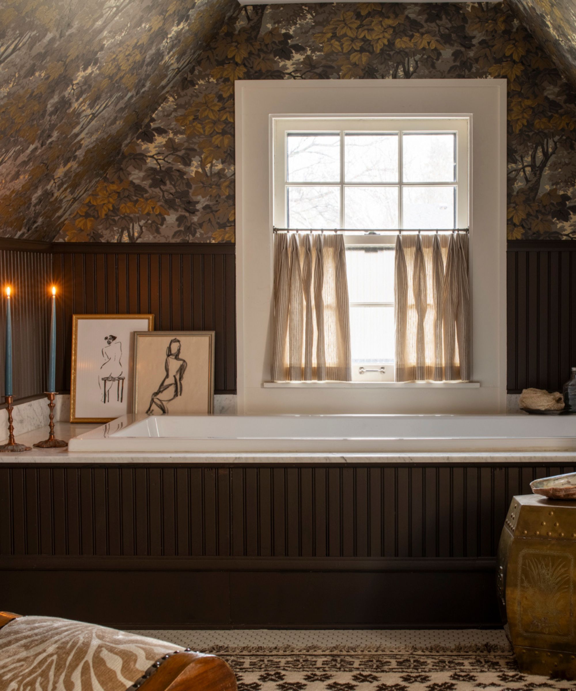 moody loft guest bathroom with dark wooden brown panelling and bath panel, small window with cafe curtain, botanical wallpaper, decorated with a rug, stool, artwork, candles and a small chair