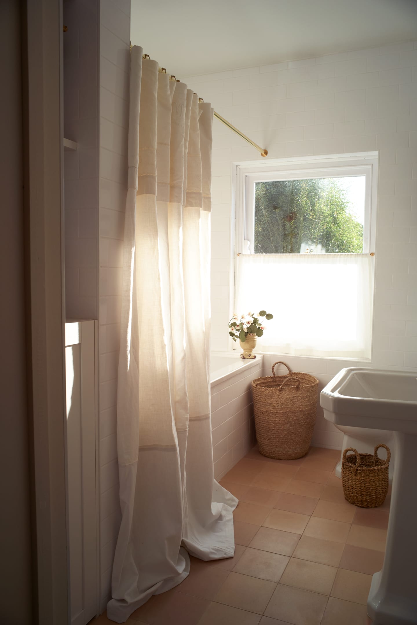 White bathroom with linen shower curtain, basket storage and terracotta floor