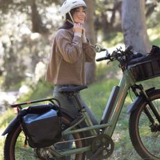 A woman doing up her helmet while stood next to a green e-bike in a wooded area