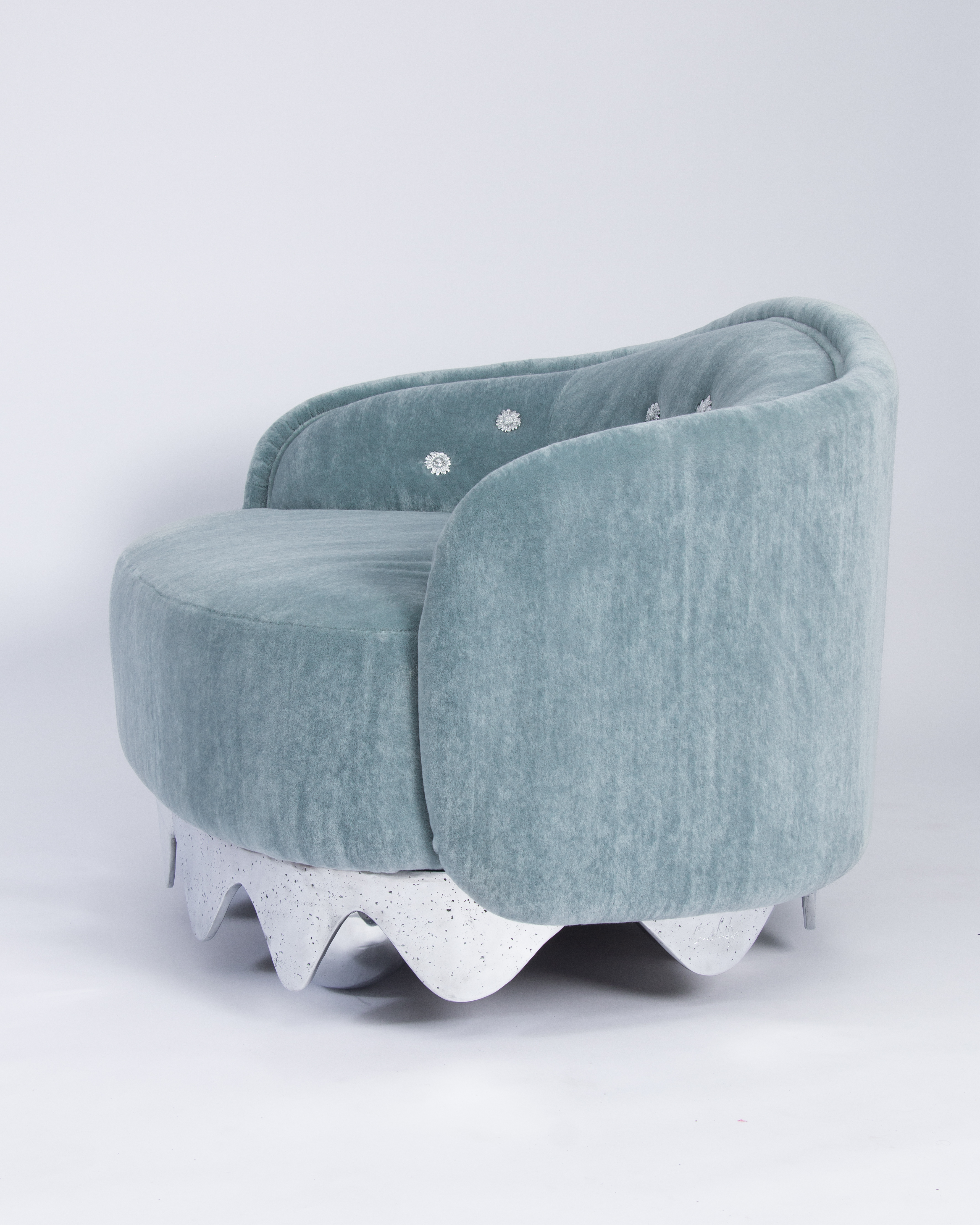 A creaturesque armchair made of pale blue velvet, a wavy chrome base, emboridered white flowers, and a soft silhouette.