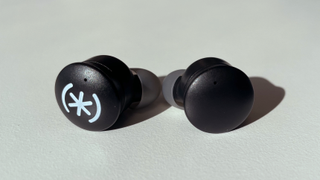 The Speck Gemtones Play earbuds sitting on a white table top. They are matte black with the Speck logo printed on the top of the left earbud in white.