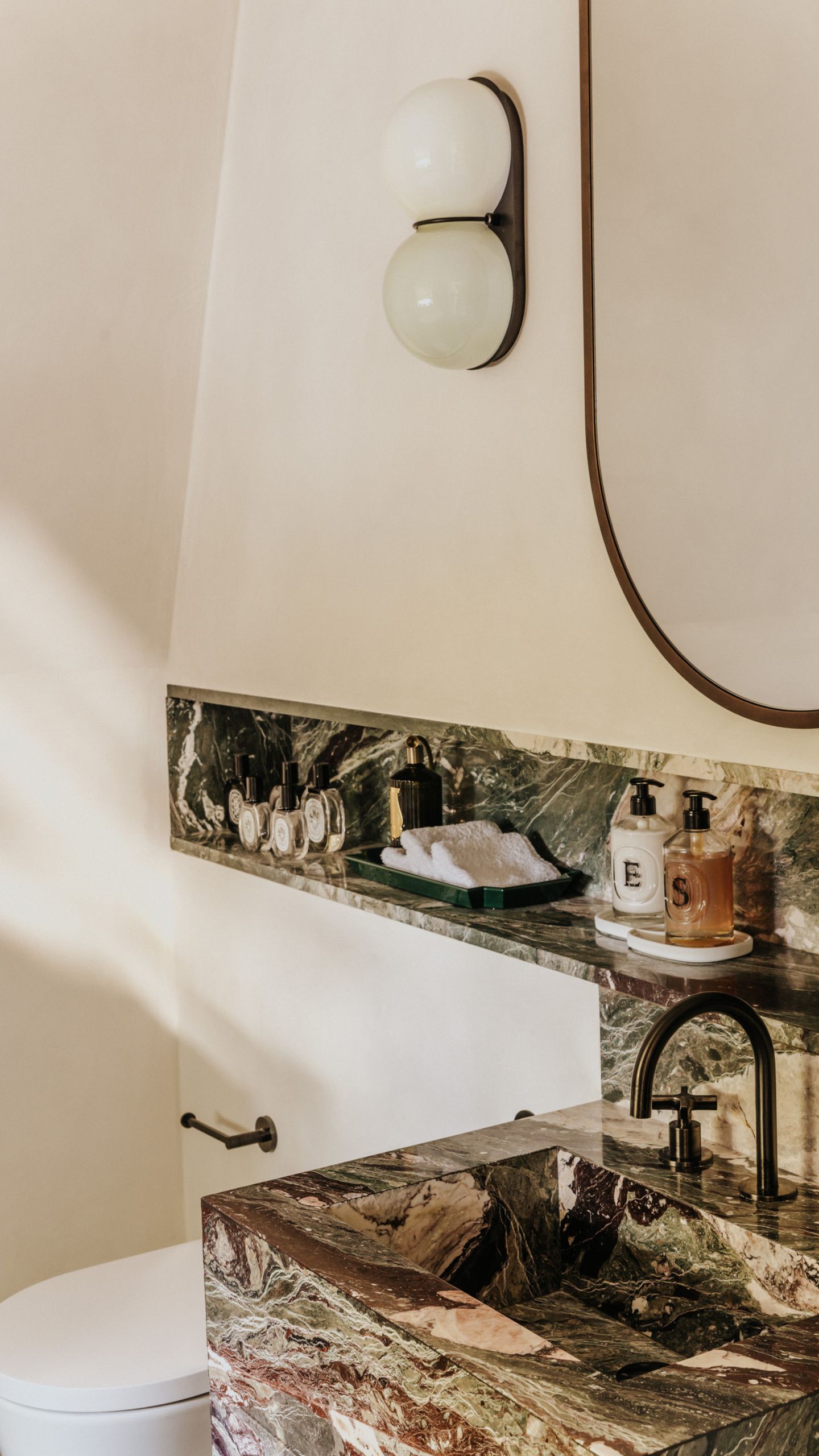 Close-up of a bathroom countertop that's made of dark green marble in a white bathroom. There is a bubble sconce light and lots of fancy soaps and room sprays.