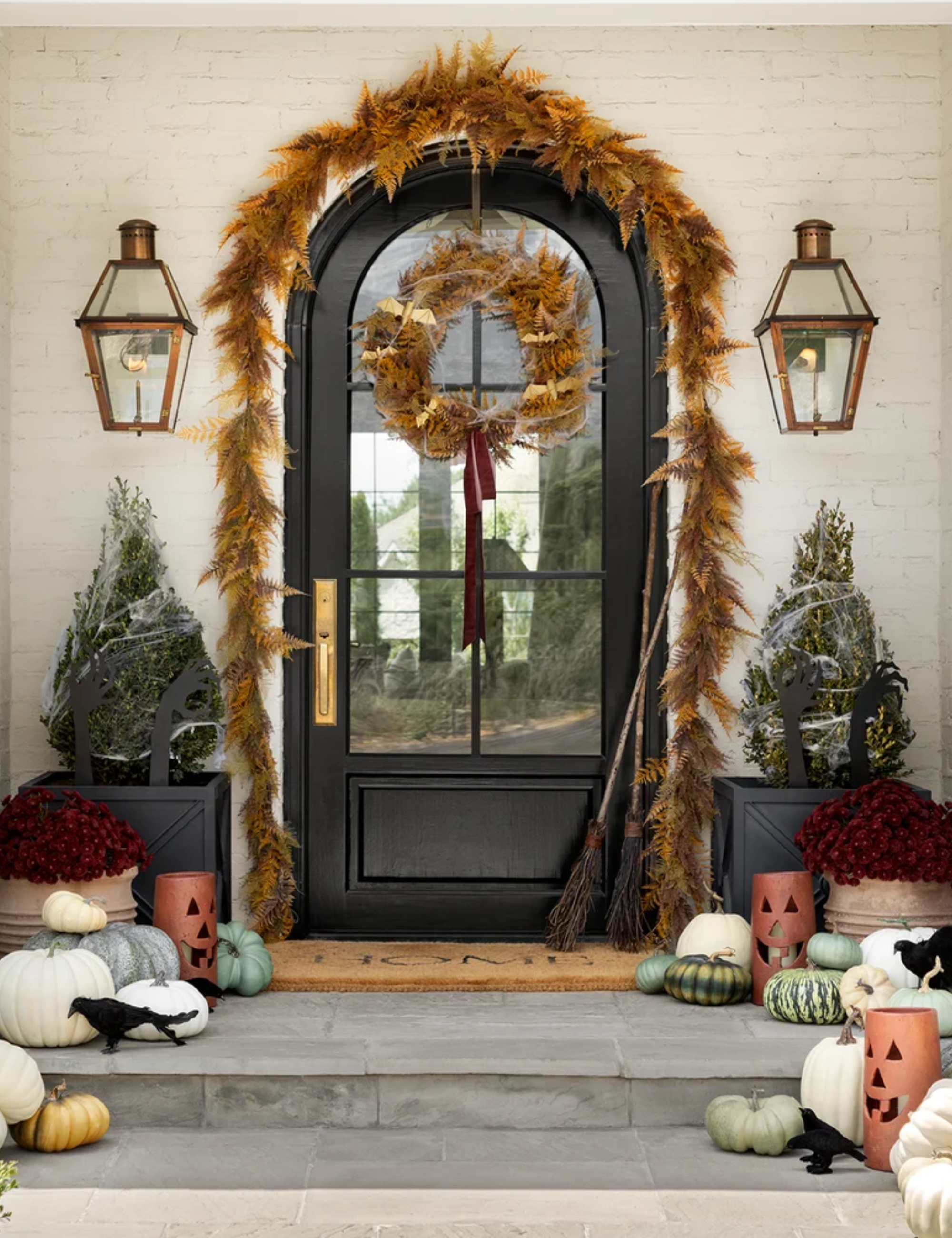 Festive Halloween porch decorated with aged fern garland, jack-o-lanterns, woven broomsticks, and scattered pumpkins