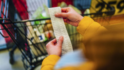 A person checking the receipt for their grocery shop, with a full shopping cart in the background.
