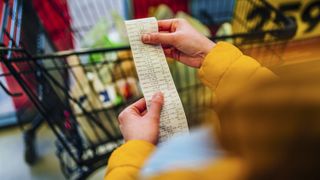 A person checking the receipt for their grocery shop, with a full shopping cart in the background.