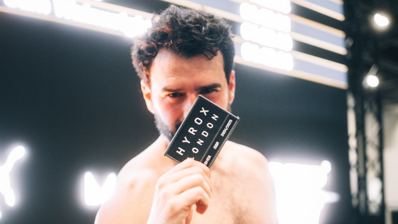 A man holds up a black patch reading &quot;HYROX London&quot; to the camera, obscuring his face