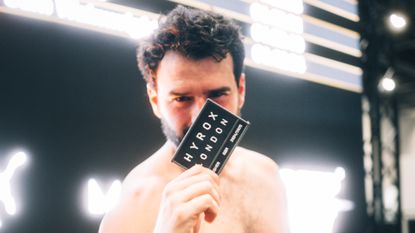 A man holds up a black patch reading "HYROX London" to the camera, obscuring his face