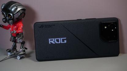 The Asus ROG Phone 8 on a grey and pink background, sat alongside some ROG merchandise