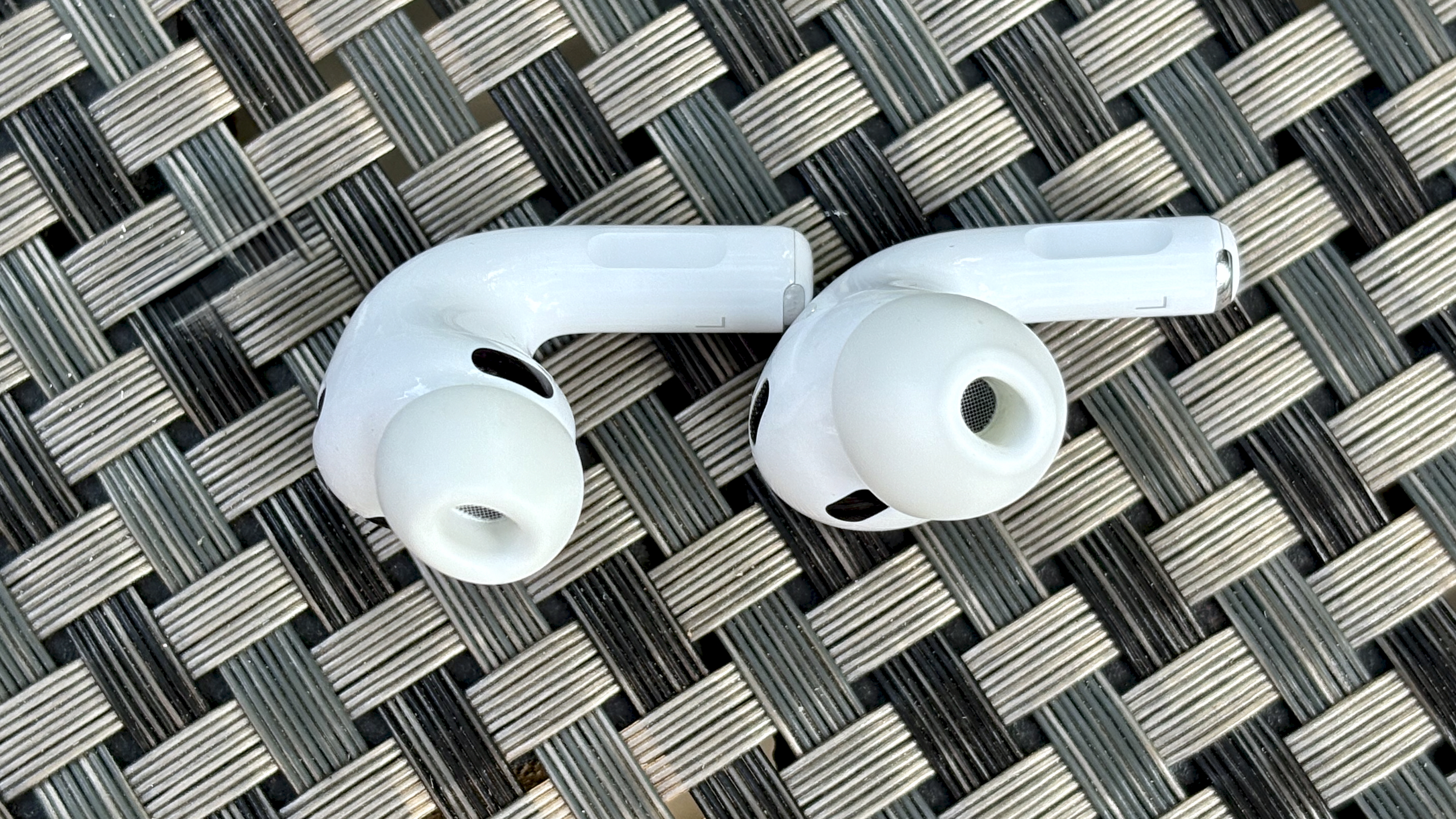 Apple AirPods Pro 3 either worn by a man standing in front of a red-brick wall, or on a table with gray twine cover 