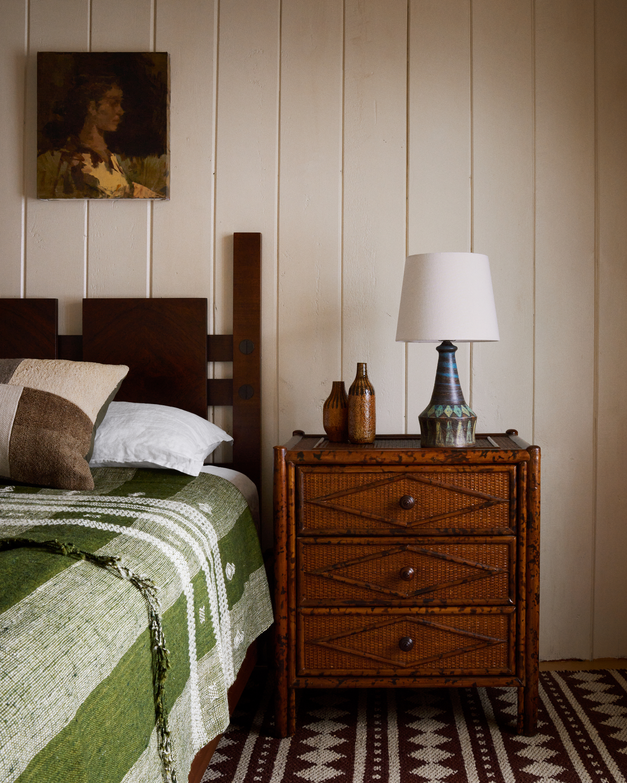 A wooden bed with green and white bedding in a white wood-panelled room. There is a white and brown rug and a wooden side table with a lamp on it.