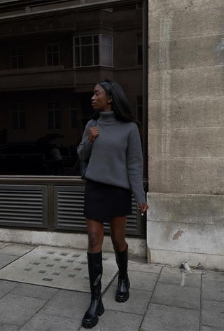 how to wear Chelsea boots is shown in a photo of a woman on the sidewalk wearing a gray turtleneck sweater with a black shoulder bag, black mini skirt, and black knee-high Chelsea boots